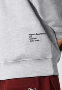 Sudadera gris con puños y bajo acanalados, que presenta un texto negro en la manga: "French Sportswear est. mil novecientos treinta y tres." Pantalones rojos son parcialmente visibles.