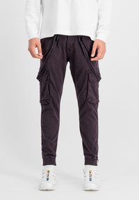 Chino-Cargohosen in dunkelviolett, mit mehreren Seitentaschen, verstellbaren Cargoriemen und elastischen Bündchen. Getragen mit einem weißen Langarmshirt.
