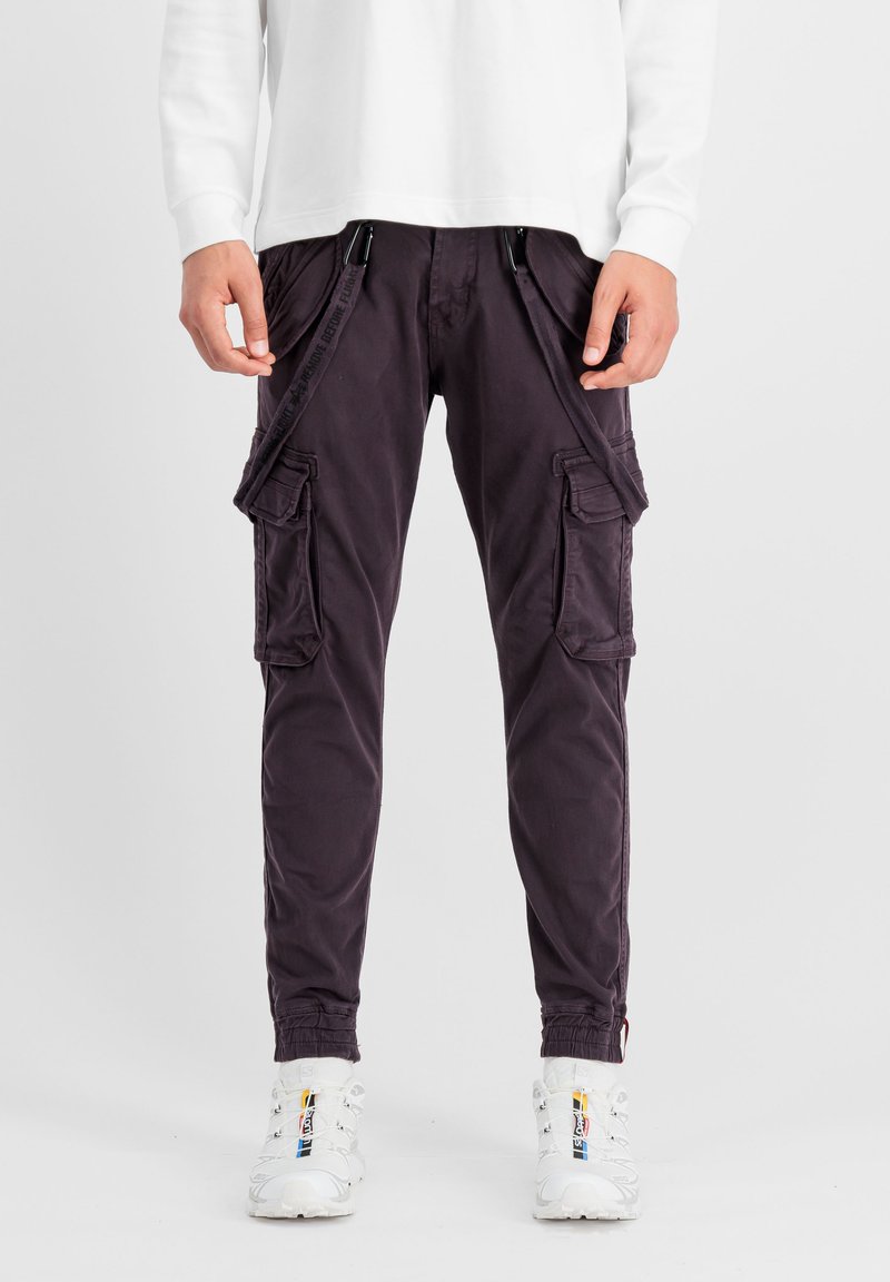 Chino-Cargohosen in dunkelviolett, mit mehreren Seitentaschen, verstellbaren Cargoriemen und elastischen Bündchen. Getragen mit einem weißen Langarmshirt.