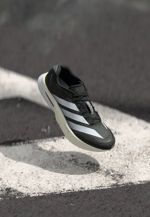 Chaussure de course Adidas Adizero noire avec des bandes blanches et une semelle blanche, semblant flotter au-dessus d'une surface asphaltée marquée.