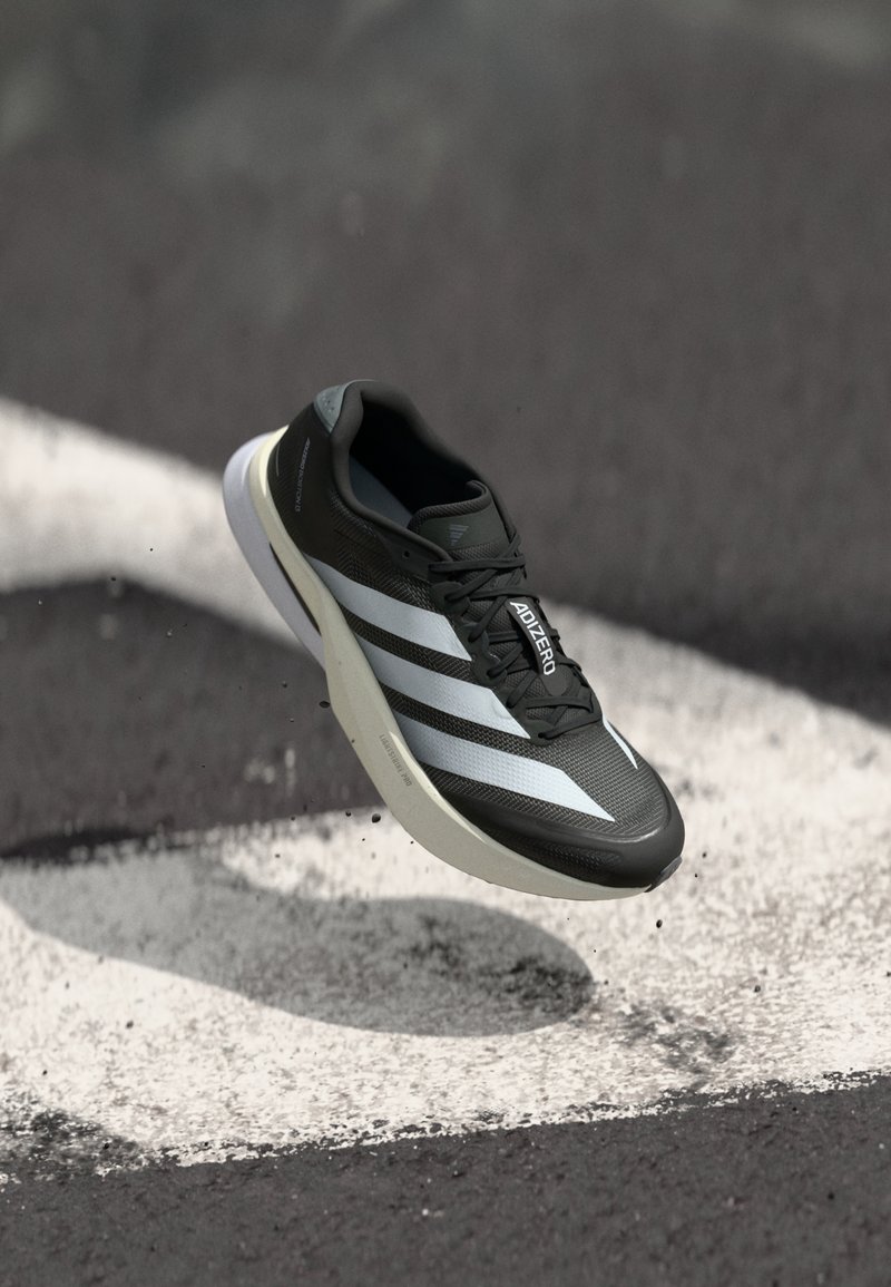 Chaussure de course Adidas Adizero noire avec des bandes blanches et une semelle blanche, semblant flotter au-dessus d'une surface asphaltée marquée.