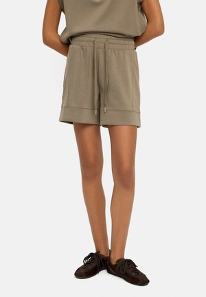 Osoba ubrana w luźne oliwkowozielone spodenki ze sznurkiem i ciemnobrązowe buty sznurowane, stojąca z ramionami za plecami.