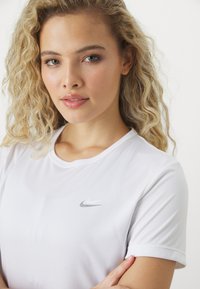 Camiseta deportiva blanca de manga corta, con cuello redondo y un pequeño logo gris de Nike en el lado izquierdo del pecho, hecha de un material suave y ligero.