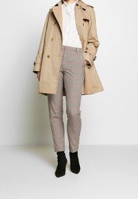 Trench beige sur une blouse blanche, associé à un pantalon à carreaux rouge, noir et blanc. Des bottines noires complètent la tenue.