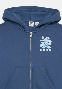 Blå hettejakke med glidelås, frontlommer og en lys blå blomster- og bølgelogo med "ROXY"-tekst på venstre bryst.