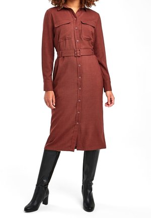 Femme portant une robe couleur rouille à manches longues, boutonnée, ceinturée à la taille, avec des poches devant et des bottes noires à talons jusqu’aux genoux, debout sur fond blanc.