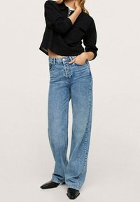 Femme portant une veste courte noire, un haut à rayures, un jean bleu taille haute à jambes larges et des chaussures noires à bout pointu, debout avec une main dans la poche.