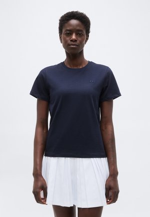 Γυναίκα όρθια, φοράει μπλε σκούρο κοντομάνικο μπλουζάκι και λευκή πλισέ φούστα, μπροστά σε απλό ανοιχτό γκρι φόντο.