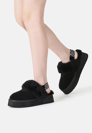 Black suede platform shoes with a fluffy black fur trim and a patterned elastic strap at the heel. Soft texture and rounded shape.