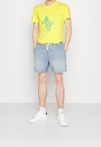 Keltainen lyhythihainen T-paita, jossa on vihreä "Polo" -grafiikka, yhdistettynä vaaleansinisiin farkkushortseihin ja valkoisiin lenkkitossuihin. Rentoa ilmettä narualla.