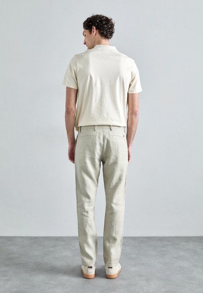 Chemise crème unie à manches courtes, pantalon en lin beige clair et chaussures beiges avec une semelle texturée, vues de dos sur fond gris.