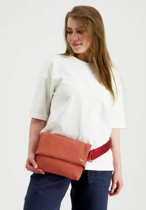 Pitkä hiuksinen nainen, jolla on valkoinen paita ja laivastonsiniset housut, pitää oranssia taitettavaa laukkua, jossa on punainen hihna vyötäröllään.