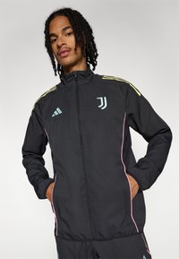 Veste zippée noire avec des accents bleu clair et rose, arborant les logos Adidas et Juventus, en matériau lisse et léger.