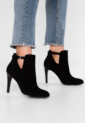 Bottines noires en daim à talons hauts avec découpe latérale et bride à boucle, portées avec un jean bleu à ourlet effiloché.