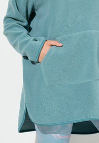 Fleece pullover pehmeas teal värvuses, millel on tekstureeritud pind, suure eesmise taskuga ja kontrastsete äärtega.