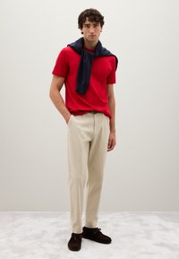 Uomo in piedi con le mani in tasca, indossa una maglietta rossa, pantaloni beige, scarpe marroni scure e un maglione blu navy drappeggiato sulle spalle, su uno sfondo neutro.