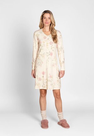 Vrouw staat en draagt een crèmekleurige, bloemenprint, nachthemd met lange mouwen, beige sokken en roze pluizige pantoffels op een effen lichte achtergrond.