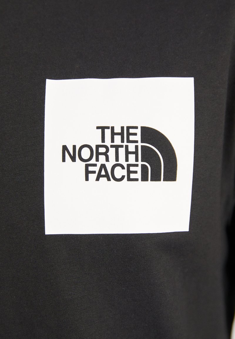 Czarna koszulka z tkaniny z białym kwadratem, w którym znajduje się logo "THE NORTH FACE" w grubym, czarnym kroju pisma oraz zakrzywionym elementem graficznym.
