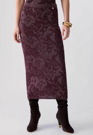 Donna che indossa una gonna midi color viola scuro con motivo floreale in tessuto strutturato e stivali neri in camoscio con tacco alto, braccialetti dorati al polso.