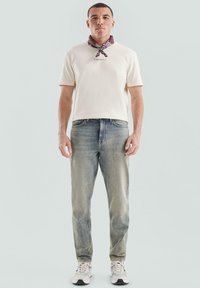 Man in een witte kortarmige shirt, vervaagde blauwe jeans, witte sneakers en een paarse bandana om zijn nek, tegen een effen achtergrond.