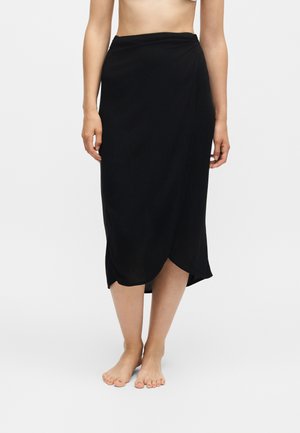 Femme portant une jupe midi noire de style portefeuille, debout pieds nus sur un fond clair uni.