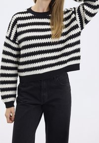 Maglione a righe in bianco e nero realizzato in maglia, con collo a girocollo e polsini a costine. Vestibilità ampia e crop. Abbinato a jeans neri a vita alta.