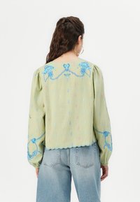 Ljusgrön blus med långa, puffärmar, scalloped nederkant och blå blommönster. Har färgglada vertikala mönster över hela.
