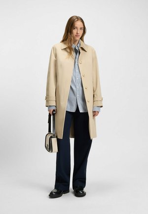 Vaaleanbeige trench-coat, jossa on klassinen kaulus ja nappikiinnitys, kerroksittain sinivalkoisen raidallisen paidan kanssa. Yhdistetty tummansinsisiin housuihin ja pieneen käsilaukkuun.