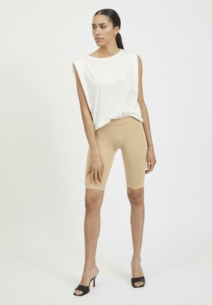 Beige biker shorts paired with a loose white sleeveless top. Model wearing black open-toe heels. Smooth fabric, casual design.