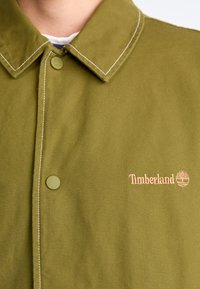 Veste olive en vert avec un col boutonné, arborant des coutures blanches et un petit logo Timberland brodé. Confectionnée en tissu durable.