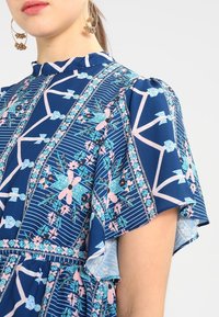 Žena v modrých šatech s růžovými a tyrkysovými květinovými a geometrickými vzory, nafouknutými krátkými rukávy a visícími zlatými náušnicemi.