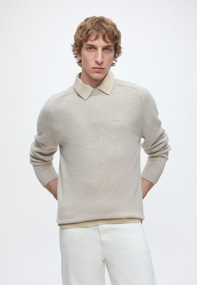 Jeune homme aux cheveux blonds bouclés portant un pull beige par-dessus une chemise à col et un pantalon blanc, debout les mains derrière le dos.