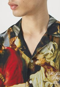 Camisa de manga corta con un diseño artístico vibrante, predominantemente negra con detalles multicolores, cuello con muesca y botones blancos.