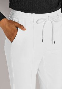Hand mit dunklem Hautton in der Tasche einer weißen Hose mit Kordelzug, kombiniert mit einem schwarzen strukturierten Oberteil.