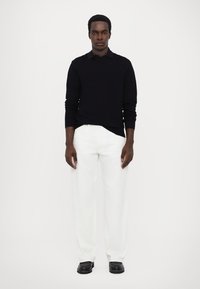 Pull en tricot bleu marine, pantalon large blanc et chaussures noires. Le modèle se tient contre un fond simple, montrant un look épuré et minimaliste.