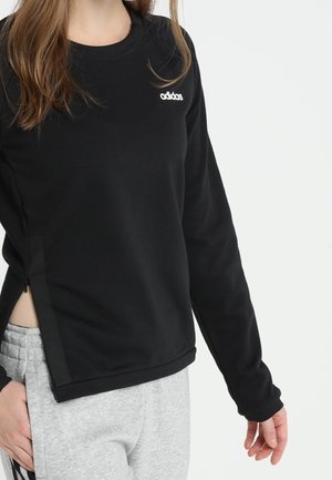 Kobieta ubrana w czarną bluzę Adidas z logo na piersi i szare spodnie dresowe, stojąca na białym tle, z rękami swobodnie opuszczonymi wzdłuż ciała.