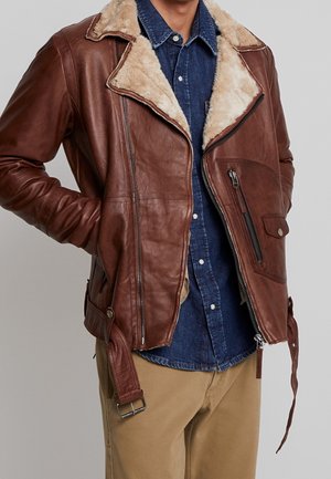 Veste en cuir marron avec col en peau lainée portée sur une chemise bleue à boutons et un pantalon beige, les mains dans les poches de la veste.