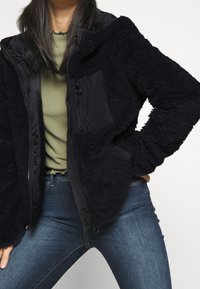 Svart fleecejacka med en mjuk textur, långa ärmar och dragkedja framtill. Har en kontrasterande slät tygpanel på bröstet.