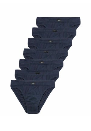 Muskelbepackter Mann mit kurzen Haaren, der dunkelblaue Slips trägt, steht vor einem schlichten hellgrauen Hintergrund und schaut zur Seite.