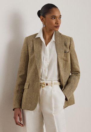 Žena v béžovom blejzri s rybej kosťou, bielej košeli na gombíky, bielych nohaviciach s vysokým pásom a opaskom z lana, sogeometrickými náušnicami, pozerá na pravú stranu.