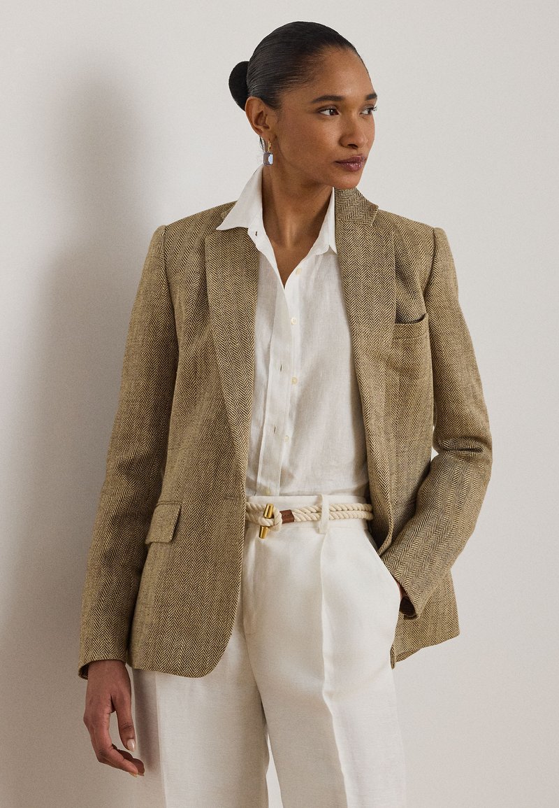 Γυναίκα που φοράει ένα μπεζ σακάκι με σχέδιο ψαροκόκαλο, λευκό πουκάμισο με κουμπιά, λευκό παντελόνι ψηλής μέσης με ζώνη από σχοινί και γεωμετρικά σκουλαρίκια, κοιτώντας δεξιά.