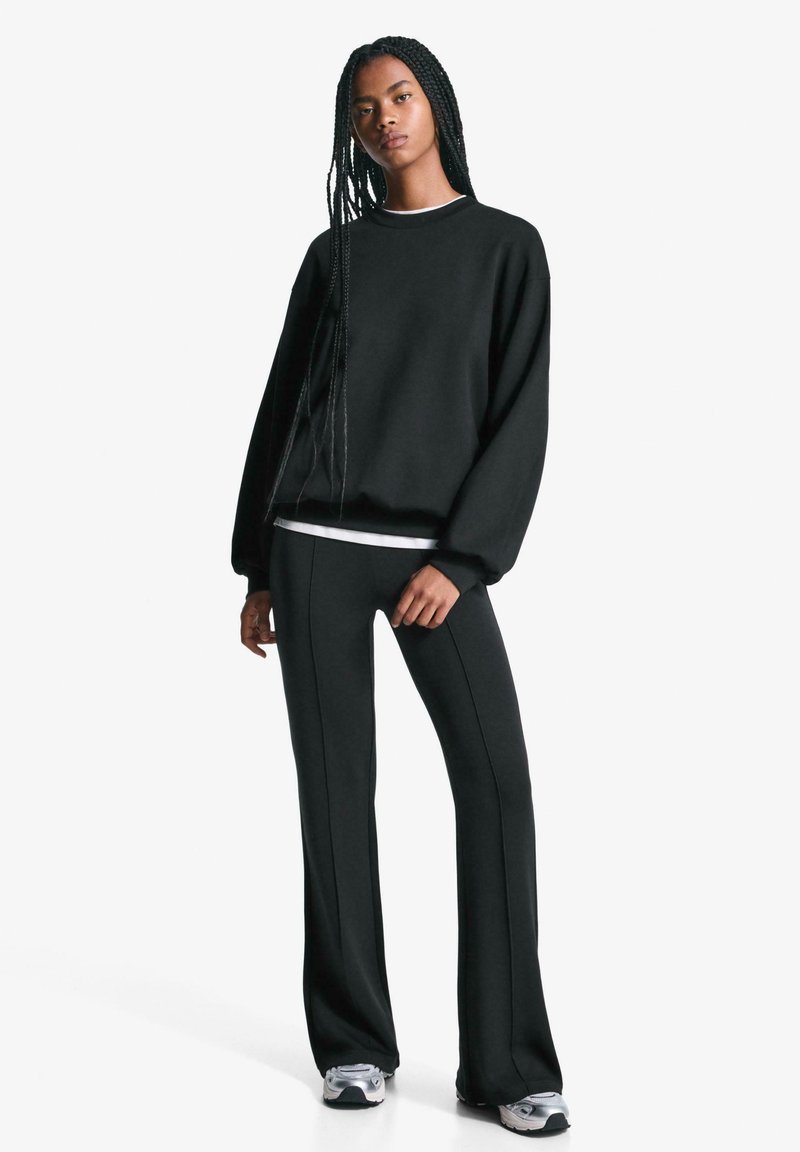 Sort sweatshirt med rund hals og dropped shoulders, parret med sorte flared bukser med sidefuger. Modellen har sportssko på.