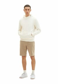 Weißer Kapuzenpullover mit Fronttasche, kombiniert mit beigen Shorts. Das Model trägt weiße Sneaker mit hellen Socken. Schlichtes Design.