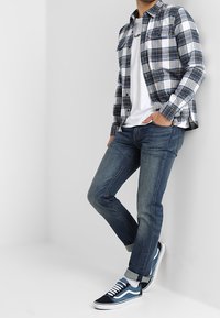 Kostkovaná košile s knoflíky v modré, bílé a šedé barvě, přes bílé tričko. UPnuté tmavé džíny a modro-bílé tenisky. Neformální outfit.