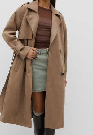 Vrouw draagt een beige trenchcoat, een bruin doorschijnend topje, een lichtblauwe denim rok en zwarte kniehoge laarzen tegen een effen grijze achtergrond.