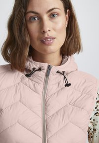 Gilet matelassé rose clair avec un col montant, fermeture éclair avant et cordons de serrage ajustables. Motif en chevrons avec une texture lisse.