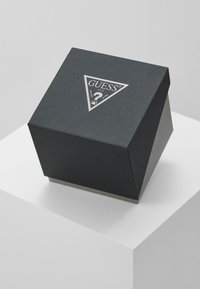 Cutie neagră cu textură triangulară, având un logo argintiu "GUESS". Cutia are un design elegant, cu un luciu discret și o bază contrastantă.
