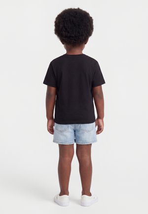 Barn med krøllet hår står med ryggen til iført en sort T-shirt, lyse denimshorts og hvide sneakers på en ensfarvet baggrund.
