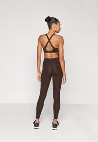 Καφέ αθλητικό στήθος με σταυρωτούς ιμάντες και λεπτομέρειες από δίχτυ, συνδυασμένο με καφέ κολάν με πλαϊνές περικοπές, φορεμένο με μαύρα αθλητικά παπούτσια.