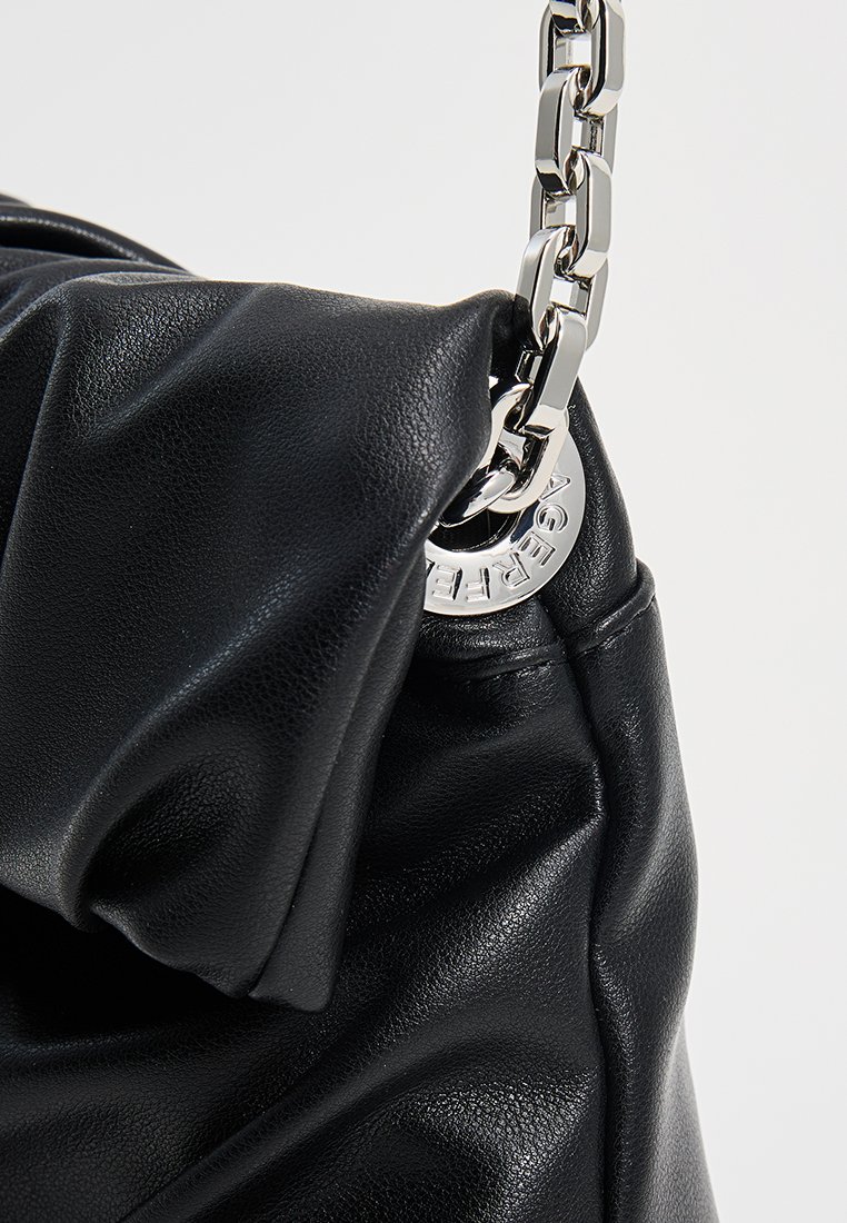 Bolso de mano de piel negra con textura fruncida, que presenta un asa de cadena plateada y un acento metálico circular grabado con el nombre de la marca.